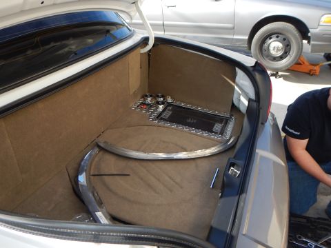 2003 Ford Crown Victoria - carpeted trunk of a sedan stands open showing a chrome amplifier and metal trim pieces, with another sedan on a jack in the background.
