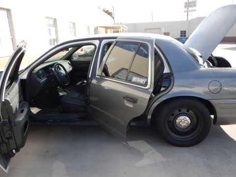 2003 Ford Crown Victoria - gray sedan sits with both driver-side doors open and the hood raised, showing its cloth interior and black steel wheels in a shop lot.