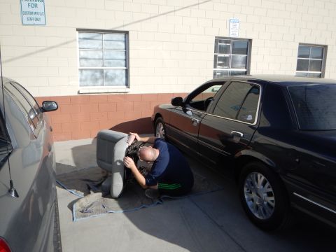 2003 Ford Crown Victoria - man kneels beside a dark sedan in a parking spot, working on a car seat resting on the ground next to a brick building.