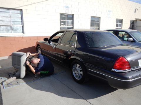 2003 Ford Crown Victoria - Rear three-quarter view of a dark full-size sedan with chrome wheels parked by a brick building, a man crouched working near its front.