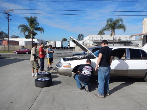 2003 Ford Crown Victoria - group of men work around a white sedan with its hood up in a sunny lot, with a chrome wheel and tire resting on the pavement.
