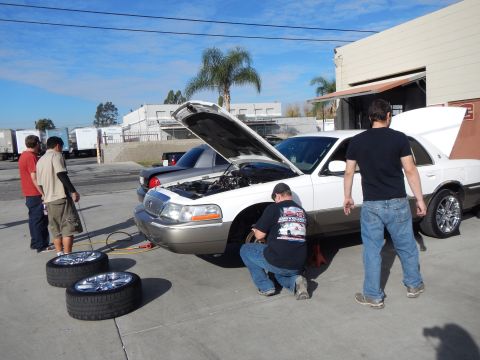 2003 Ford Crown Victoria - Men gather around a white sedan with its hood raised in a shop lot, two chrome wheels on the ground and box trucks in the background.