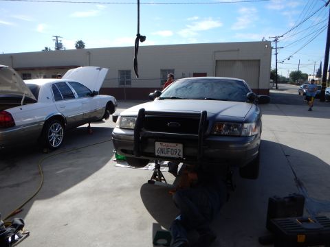 2003 Ford Crown Victoria - gray sedan fitted with a black front push bar sits jacked up with a person working underneath; its plate reads 6NUF092.