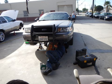 2003 Ford Crown Victoria - Front view of a gray Ford sedan with a black push bar, license plate 6NUF092, as a man lies beneath it working on the front end.