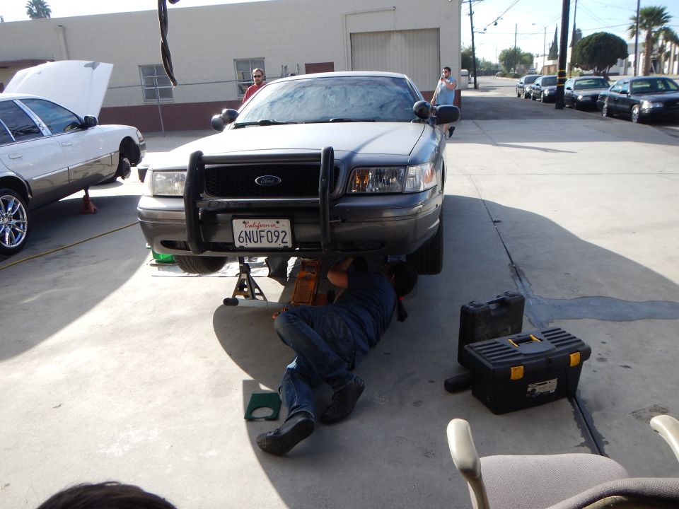 Front view of gray Ford sedan with push bar, plate 6NUF092, man lying underneath working