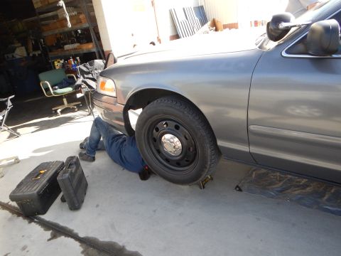 2003 Ford Crown Victoria - person lies under the front corner of a gray sedan with the steel wheel removed, a toolbox beside them in a cluttered garage.