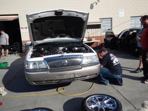 2003 Ford Crown Victoria - silver Mercury sedan sits with its hood raised as a man crouches at the front grille, a chrome wheel resting in the foreground.