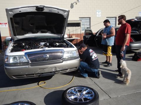 2003 Ford Crown Victoria - Men and a small dog stand around a silver Mercury sedan with its hood up while one crouches at the grille, a chrome wheel in the foreground.