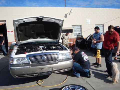 2003 Ford Crown Victoria - silver Mercury sedan stands with its hood raised in a shop lot surrounded by people, with a chrome wheel resting in the foreground.