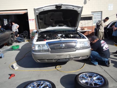 2003 Ford Crown Victoria - silver Mercury sedan sits with its hood open as a man crouches at the front, two chrome wheels lying on the pavement nearby.