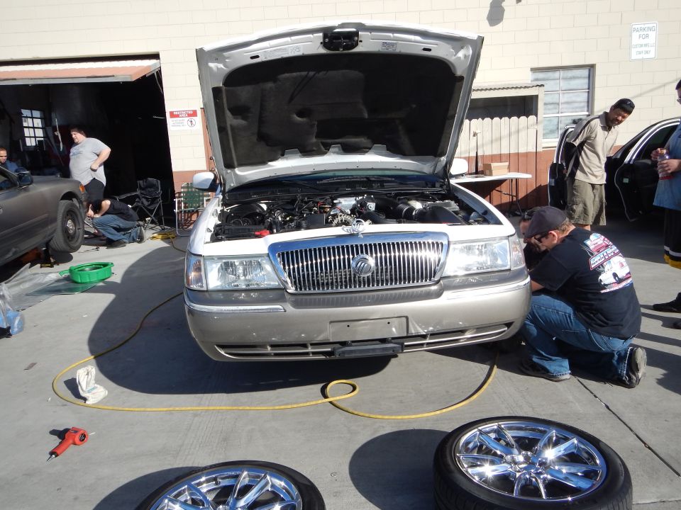 Silver Mercury sedan hood up, man crouched at front, two chrome wheels on the ground
