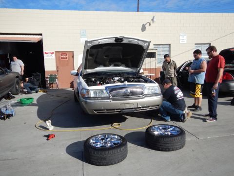 2003 Ford Crown Victoria - Men gather around a silver sedan with its hood raised in a sunny shop lot, with two large chrome wheels and tires on the pavement in front.