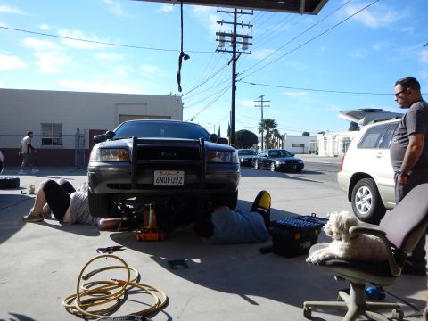 2003 Ford Crown Victoria - gray Ford sedan with a black push bar sits jacked up with two people working beneath it, a small dog resting on a chair nearby.