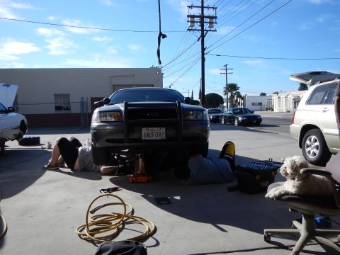 2003 Ford Crown Victoria - Front view of a gray Ford sedan with a black push bar and plate 6NUF092, two people lying underneath working in a sunny shop lot.