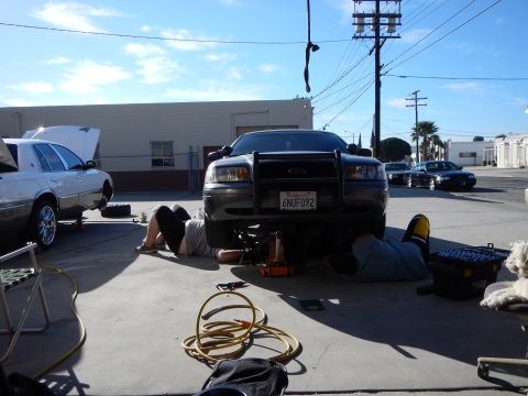 2003 Ford Crown Victoria - gray Ford sedan with a black push bar sits raised with plate 6NUF092 as two people work beneath it, a white sedan and hose in the lot.