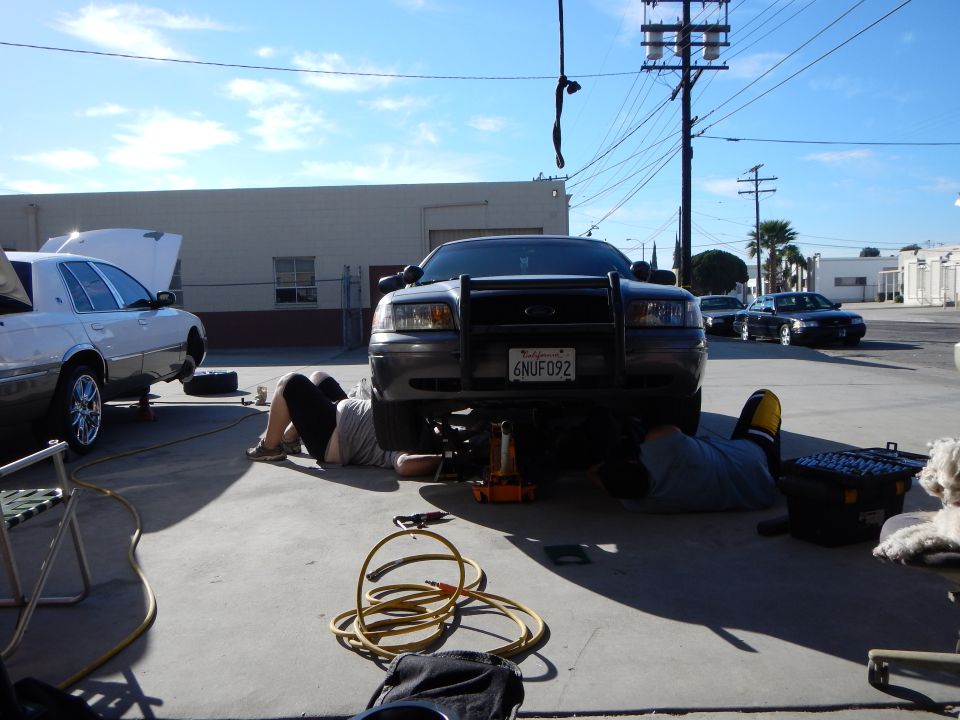 Gray Ford sedan with push bar lifted, plate 6NUF092, two people working under it