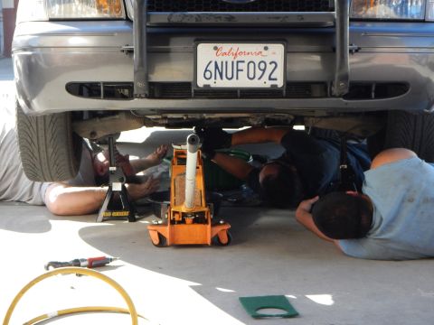 2003 Ford Crown Victoria - Three people work underneath the front end of a silver vehicle raised on jack stands, with an orange floor jack and tools on the ground.