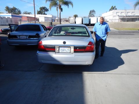 2003 Ford Crown Victoria - Rear view of a silver Mercury sedan in a lot, a bald man in a blue shirt standing beside it, with another sedan nearby.