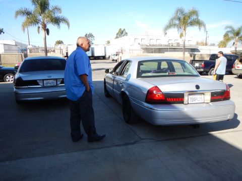 2003 Ford Crown Victoria - man in a blue shirt stands between two silver sedans parked in a sunny industrial lot with palm trees and buildings.