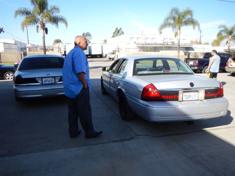 Man in blue shirt beside two silver sedans in a paved industrial lot under blue sky