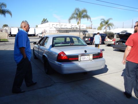 2003 Ford Crown Victoria - silver sedan seen from the rear in a paved lot, with a man in a blue shirt and several other people and vehicles around.