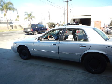 2003 Ford Crown Victoria - Driver-side profile of a silver Mercury sedan with black-painted wheels parked outside a workshop with cars and people behind.