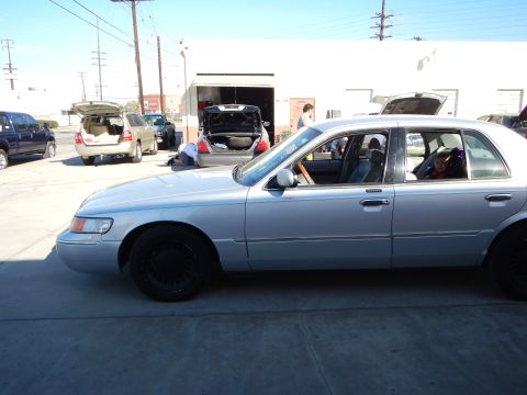 2003 Ford Crown Victoria - silver Mercury sedan with black wheels shown in side profile, with open trunks, people and other cars behind it at a shop.