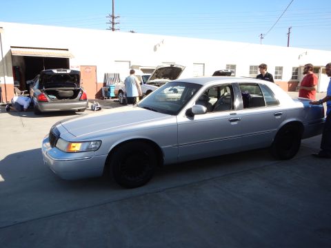 2003 Ford Crown Victoria - Front three-quarter view of a silver Mercury sedan with black wheels parked in a sunny lot, with people and cars in the background.