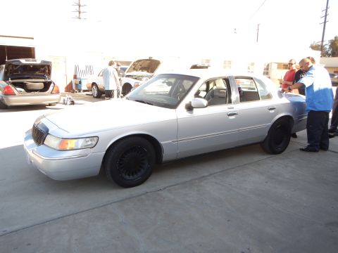 2003 Ford Crown Victoria - silver Mercury sedan with black wheels parked in a lot while two men in blue stand at its rear and other cars sit behind.