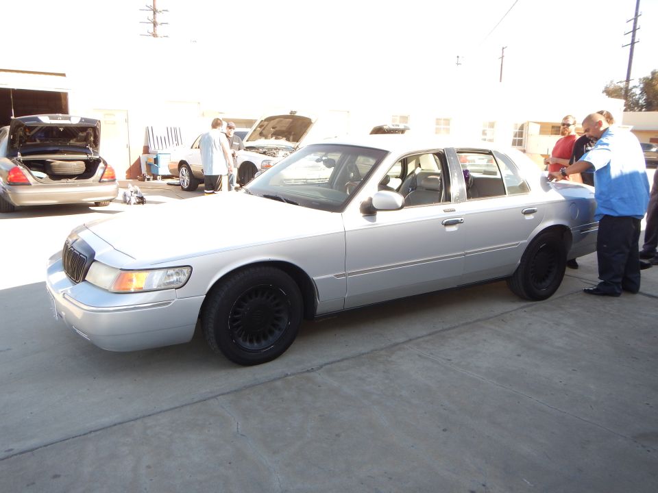 Silver Mercury sedan with black wheels, men in blue gathered beside it in a lot