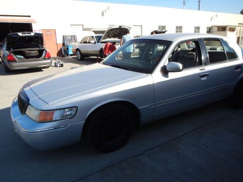 2003 Ford Crown Victoria - Front three-quarter view of a light silver Mercury sedan with black wheels parked outside a workshop with open-trunk cars behind.