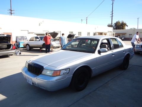 2003 Ford Crown Victoria - silver Mercury sedan with black wheels parked in a lot, with several people working on other cars in the background.