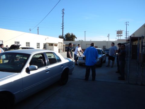 2003 Ford Crown Victoria - group of people gather and talk in a paved industrial lot with parked sedans, buildings and power poles under a clear sky.