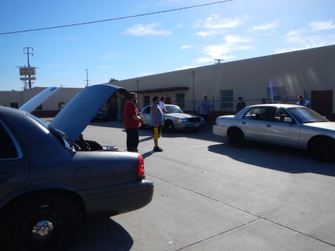 2003 Ford Crown Victoria - Several sedans parked in a sunny industrial lot, one with its hood raised, while a few people stand and talk nearby.