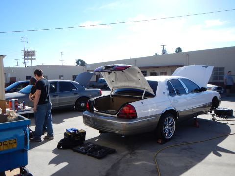 2003 Ford Crown Victoria - white sedan raised on a jack with its trunk and hood open, a person and toolboxes beside it in a workshop lot.