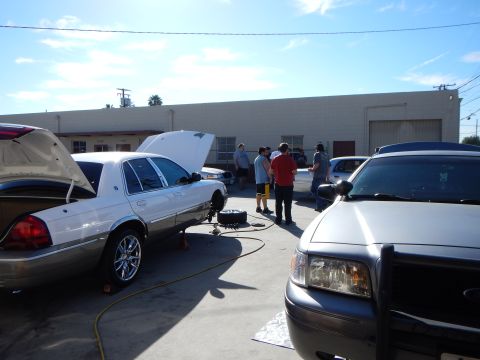 2003 Ford Crown Victoria - white sedan with its hood up sits in a lot beside a gray SUV in the foreground while people gather and work nearby.