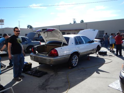 2003 Ford Crown Victoria - white sedan raised on a jack with trunk and hood open, a man in a black t-shirt nearby and a crowd of people behind.