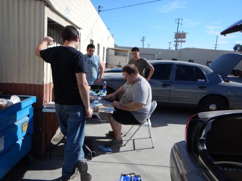 2003 Ford Crown Victoria - Several men gather around a folding table working on parts beside parked cars and a blue dumpster in a sunny lot.