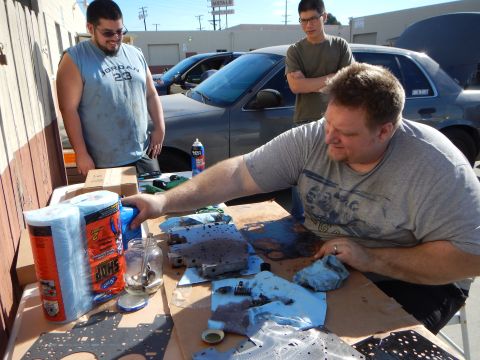 2003 Ford Crown Victoria - man works on a disassembled automatic transmission valve body spread on cardboard at a table while two men look on.