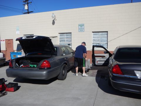 2003 Ford Crown Victoria - gray sedan with its trunk and door open in a lot, a man in a blue shirt beside it next to a dark sedan and a building.