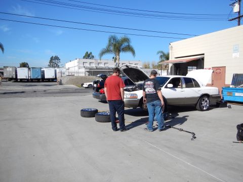 2003 Ford Crown Victoria - Two men work on a white sedan with its hood up in an industrial lot, with spare tires lying on the pavement nearby.