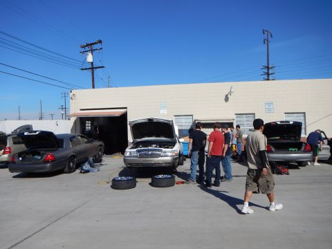 2003 Ford Crown Victoria - row of sedans with raised hoods and open trunks while several people work and gather in a sunny industrial lot.
