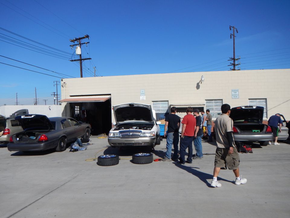 Row of sedans with open hoods and trunks, people working in a sunny industrial lot