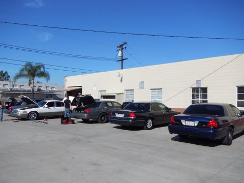 2003 Ford Crown Victoria - Several sedans parked along a beige industrial building, with a couple of people servicing one of the cars in the lot.