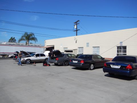 2003 Ford Crown Victoria - row of sedans parked beside an industrial building under blue sky, with people working on one car in the paved lot.