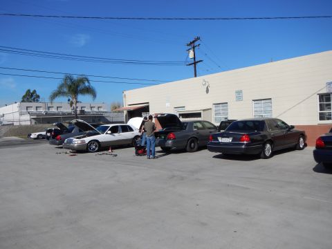 2003 Ford Crown Victoria - Multiple sedans with open hoods and trunks parked beside a building while people work between them in a sunny lot.