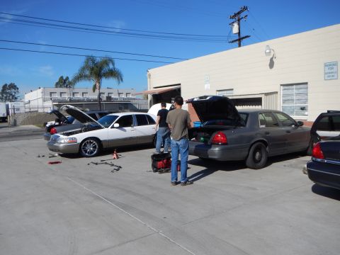 2003 Ford Crown Victoria - white Lincoln sedan and a gray Ford Crown Victoria sit with hoods open as men work on them in a sunlit industrial parking lot.
