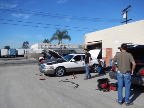 2003 Ford Crown Victoria - white Lincoln Town Car sits with its hood raised while several men work around it next to a red tool bag in an industrial lot.