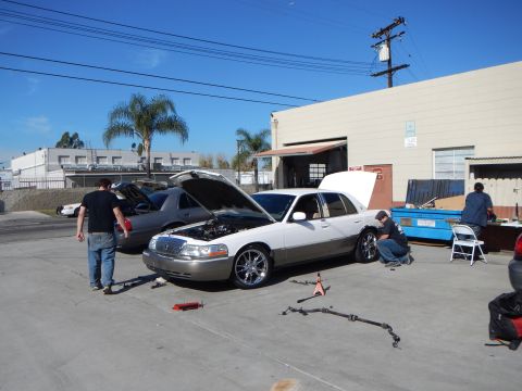 2003 Ford Crown Victoria - white Lincoln Town Car with chrome wheels sits hood-up while a man crouches by the front fender in a bright industrial lot.