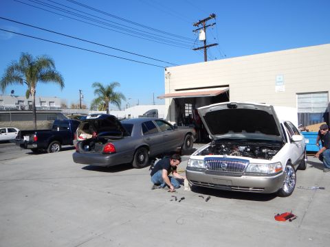 2003 Ford Crown Victoria - silver Mercury sedan with its hood up and a gray Crown Victoria with open trunk sit in a lot as a man crouches working between them.